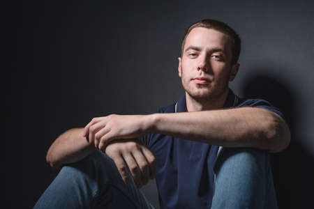Young attractive man in blue jeans and a polo shirt sits on the floor near the wall in the studio....