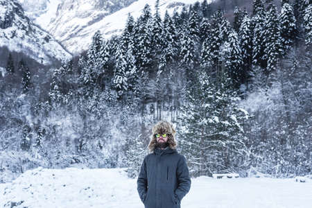An adult brutal man with a beard in ski goggles and a hat stands in the mountains in winter. Active...