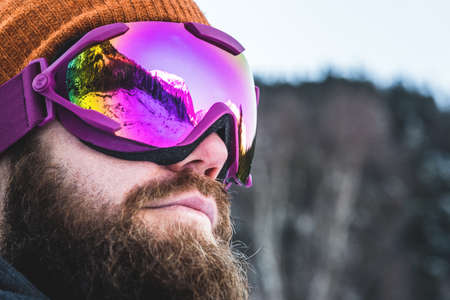 An adult brutal man with a beard in ski goggles and a hat stands in the mountains in winter. Active recreation conceptの写真素材