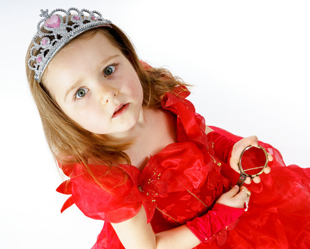 Cute little princess dressed in red looking to the compact mirror isolated on white backgroundの写真素材