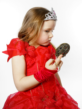 Cute little princess dressed in red looking to the compact mirror isolated on white backgroundの写真素材