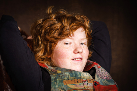 Red-haired expressive teenage boy sitting in arm-chair, studio portraitの写真素材