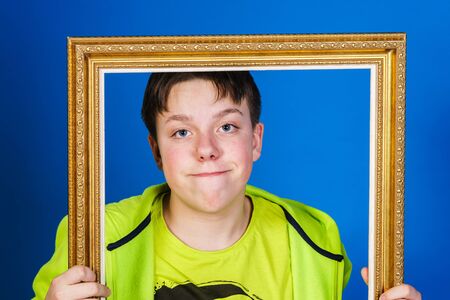 Affective teenage boy posing with picture frame, isolated on blue backgroundの写真素材
