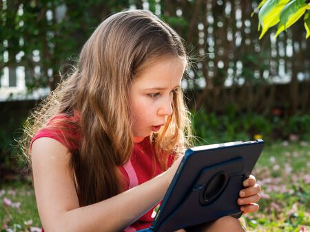 Distance learning during the quarantine period for the virus Covid-19. Cute little schoolgirl with long hair is studying from home, sitting in the garden on the grass. Uses a tablet and remote work via the Internet.の写真素材