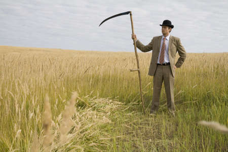 man in suit and hat with scythe standing in field with golden grassの写真素材