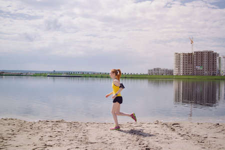 Young healthy lifestyle woman running at sunrise beach. Girl running at sand near water on city's beach. Girl running in yellow sports shirt and black shortsの写真素材