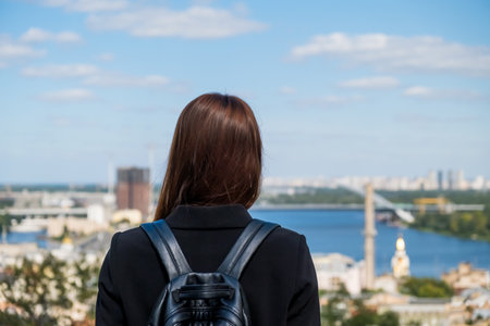 Girl looks at the city from a hill. Rear view, long hair, black backpack. Blue sky, river, crane.