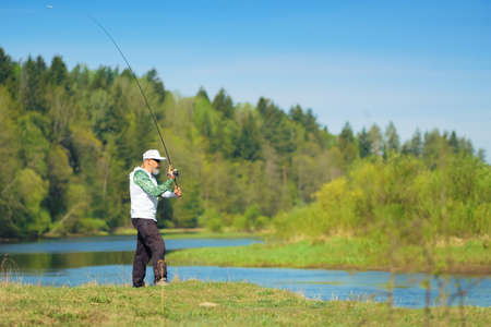 Fisherman with a spinning rod catching fish on a river at sunny summer day with green trees at background. Outdoor weekend activity. Photo with shallow depth of field taken at wide open aperture.の写真素材