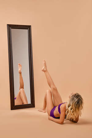 Woman with slim tanned body sits on the floor in studio in front of the mirrorの写真素材