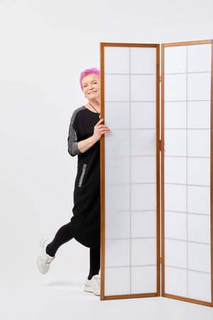 Playful old woman with short pink hair looks out from behind a screen in white roomの写真素材