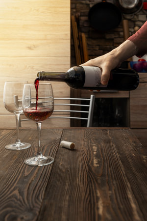 A hand pours red wine from a bottle into two elegant glasses set on a dark wooden table. The warm lighting creates a relaxed ambiance, perfect for a casual gathering or intimate dinner.の写真素材