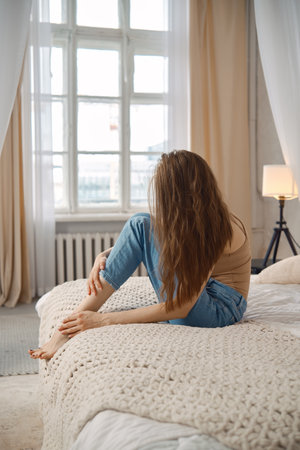 A woman with long hair is sitting cross-legged on a bed in a well-lit, serene room. Soft curtains sway gently by an open window, and a lamp provides a warm ambiance.の写真素材