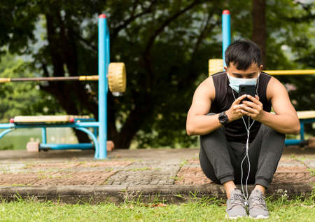Asian man sitting in a park wearing a jogging suit wearing a mask on his smartphone.の写真素材
