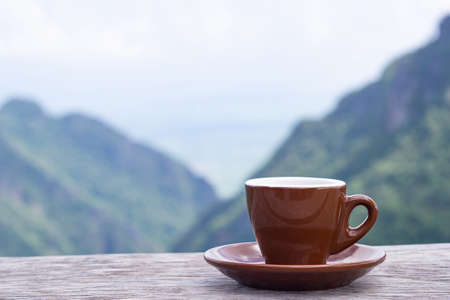 A brown coffee cup on wooden table with beautiful scenery view of mountains. Morning coffee feeling happy and freedom. Space for text.の写真素材