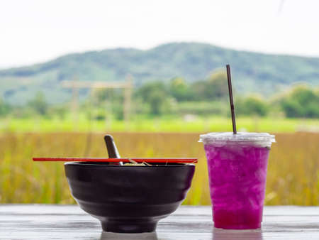 Close-up of Noodle Soup in black cup with red chopsticks and Herb water on white wooden table.