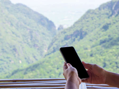 Close-up Of hands holding black smart phone on bamboo table with mountains background.の写真素材