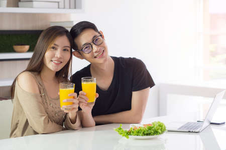 Asian young couple sitting in the kitchen and holding a glass of fresh orange juice for health with vegetables salad a laptop and smart phone placed on the table. Couple smile happy is a good healthの写真素材