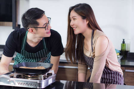 Young couple wearing apron and helping to make a steak at the kitchen in their home grill the steak on a small gas stove in the kitchen. Woman and Man smile and look steak in a pan. Couple conceptの写真素材