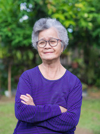 Senior woman short gray hair, wearing glasses, standing and arms crossed in her garden. Good healthy and happy life because have positive thinking. Relaxation concept.の写真素材