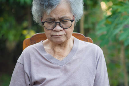 Elderly woman with short white hair, wearing glasses, eye closed and sitting on chair in the garden. Asian senior woman worried about her health problems. Health care conceptの写真素材
