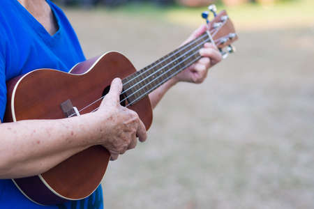 Close-up of hands elderly woman playing ukulele in her garden. Relaxing by play small guitar happy. Selective focus. Concept of old people and health careの写真素材