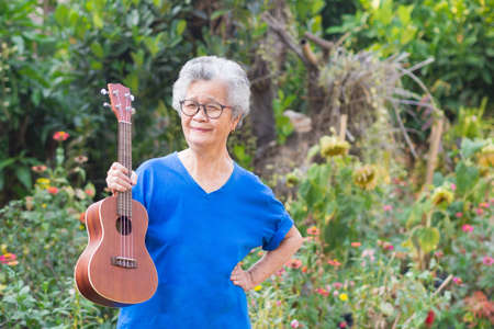 Portrait of elderly woman standing and holding ukulele in garden. Relaxing by singing and play small guitar happy and enjoy life after retired. Concept of old people and health care. Space for textの写真素材