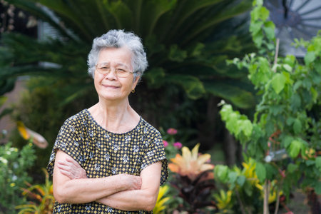 A portrait of an elderly woman short white hair wearing glasses smiling with arms crossed and looking at camera while standing in a garden. Concept of old people and health careの写真素材