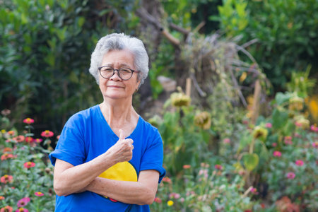 A portrait of an elderly woman short white hair wearing glasses smiling with arms crossed and showing thump up while standing in a garden. Concept of old people and health careの写真素材