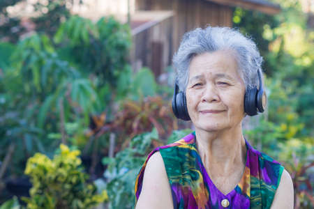 Portrait of an elderly Asian woman wearing wireless headphones listening to a favorite song and looking at the camera while standing in a garden. Concept of aged people and relaxation.の写真素材