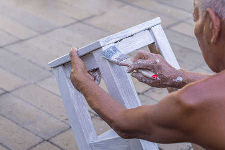 Senior man holding brush for paint white on old wooden chair without a backrest Hand of man. Old chairs are painted white. Renovation of old furnitureの写真素材