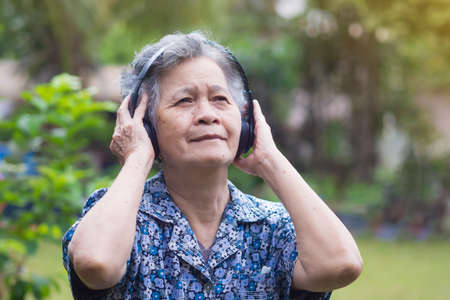 Senior Asian woman wearing wireless headphones listening to a favorite song while standing in a garden. Concept of aged people and healthcare.の写真素材