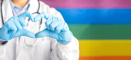 Doctor hand in blue gloves showing heart sign against the rainbow flag (LGBT) background. Medical help prophylaxis. Close-up photo. Space for text. Cardiology, healthcare and encouragement concept.の写真素材