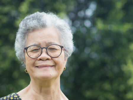 Portrait of a senior woman with short gray hair, wearing glasses, smiling, and looking at the camera while standing in a garden. Aged people and relaxation concept.の写真素材
