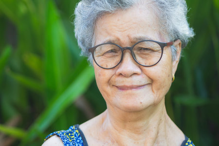 Portrait of a senior woman with short gray hair, wearing glasses, and looking at the camera with a smile while standing in a garden. Aged people and relaxation concept.の写真素材