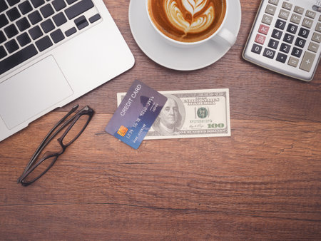 Top view of a blue credit card over on dollar banknote with a laptop, a coffee cup, calculator, and glasses on wooden table with copy space for text.の写真素材