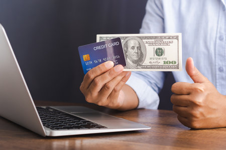 Close-up of hand holding a credit card and dollar banknotes for shopping online with a laptop. Business, e-commerce, and payment concept.の写真素材