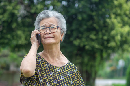 Happy beautiful senior woman is using a smartphone and looking up while standing in a garden. Space for text. Aged people and communication concept.の写真素材