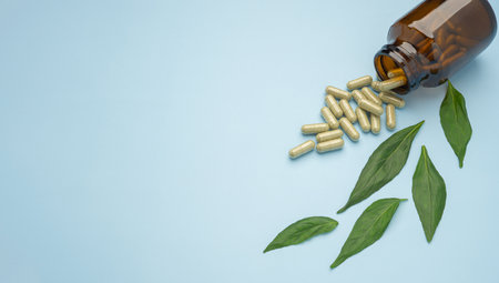 Close-up of Andrographis paniculata capsules spilling out of a drug bottle with green leaves isolated on a light blue background. Space for text. Herbal, medicine, and healthcare concept.の写真素材