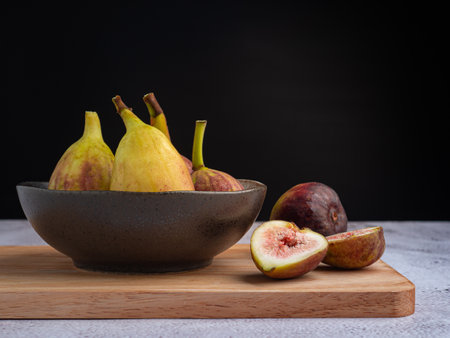 Figs fruit are in a ceramic bowl on a wooden cutting board on a cement floor with a black wall...