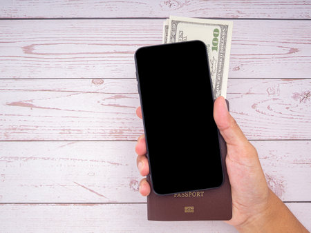 Close-up of hand holding a smartphone with passport and US dollars banknote on wooden table background. Communication and travel concept.の写真素材