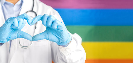 Doctor wearing gloves hands showing heart shape against the rainbow flag (LGBT) background. Medical help prophylaxis. Close-up photo. Space for text. Cardiology, healthcare and encouragement concept.の写真素材