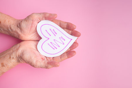 White paper cut a heart shape on the palm of a senior woman with a pink background. Healthcare, World heart day, World health day. Valentine's day. People, age, love, and healthcare concept.の写真素材