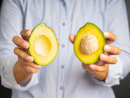 Hands holding a ripe avocado cut in half while standing in the studio. Natural fresh organic exotic fruit. Healthy eating, food diet. High Vitamin fruit. Close-up photo. Healthy fruit concept.の写真素材