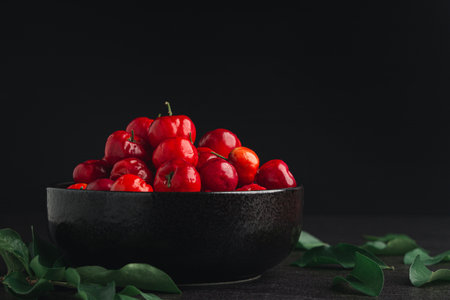 Ripe red acerola cherries fruit in a ceramic bowl and green leaves with a black background. High vitamin C and antioxidant fruits. Close-up. Space for text. Concept of healthy fruits.の写真素材