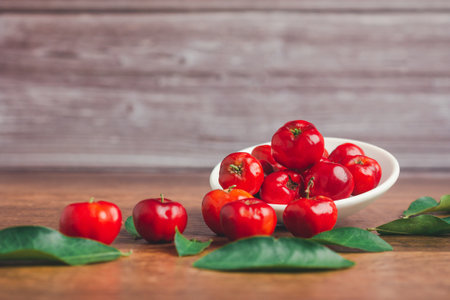 Ripe red acerola cherries fruit in a ceramic bowl and green leaves on a wooden table with a wooden...