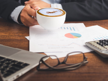 Hand of a businessman holding a coffee cup and reading financial data report while sitting in the office. Close-up photo.の写真素材