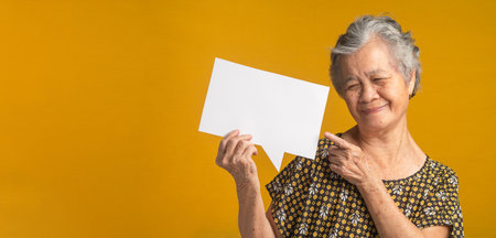Concept of a paper speech bubble. An elderly Asian woman with short gray hair holding looking and pointing at a blank paper with a smile while standing on a yellow background in the studio.の写真素材