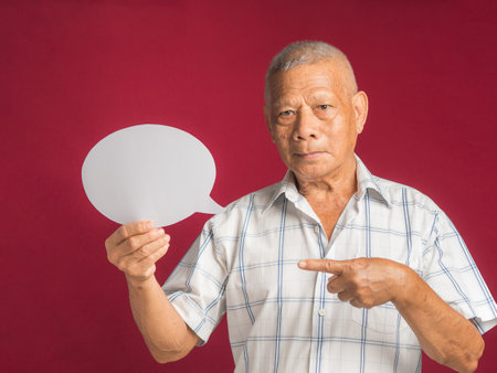 A speech bubble concept. Portrait of a senior man with short gray hair holding and pointing a blank paper speech bubble and looking at the camera while standing on a red background in the studio.の写真素材