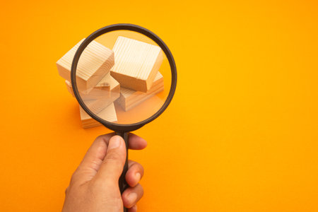 Hand holding a magnifying glass looking to wooden cubes stacked on the yellow ground. Space for text. Search concept.の写真素材