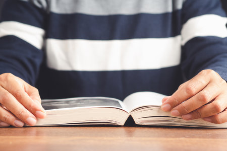 A young man in a sweater reading a book while sitting on a chair in his living room. Education,...